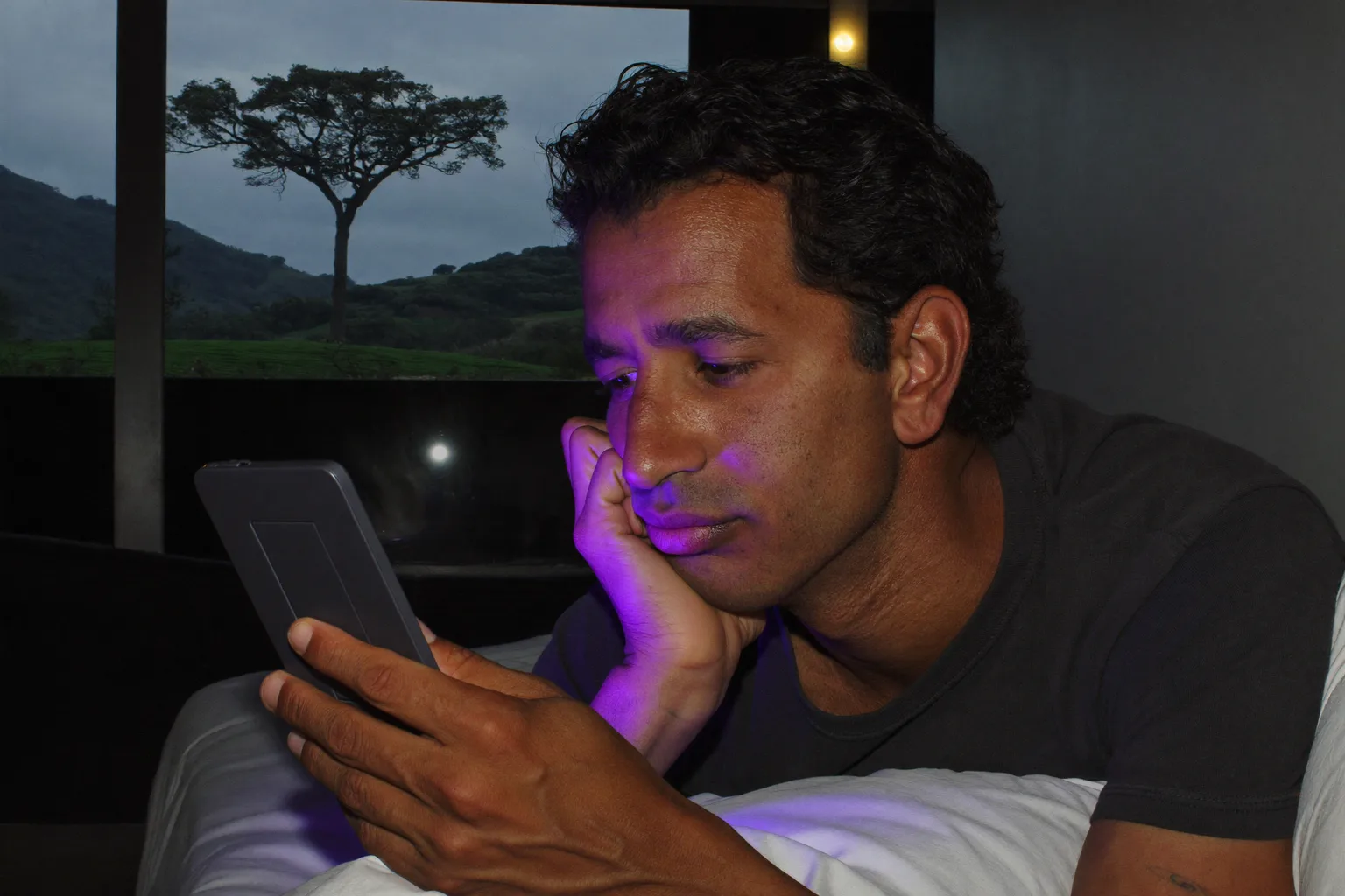 Um homem lendo em um leitor digital na cama, tarde da noite, com a página em um brilho fraco que mal alcança o rosto dele, enquanto no criado-mudo ao lado um celular virado para cima inunda a segunda cúpula do abajur com uma luz azul dura e fria