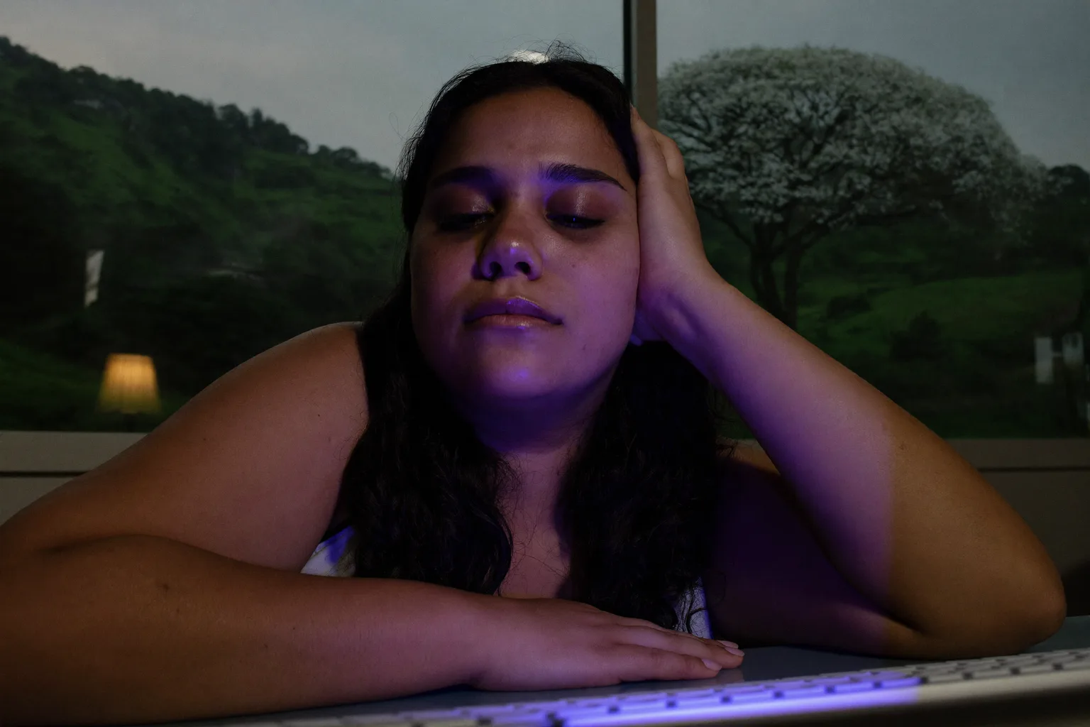Uma mulher cansada em frente ao notebook no fim do dia, com a cabeça apoiada na mão, olhos pesados e secos, olheiras, num ambiente com pouca luz