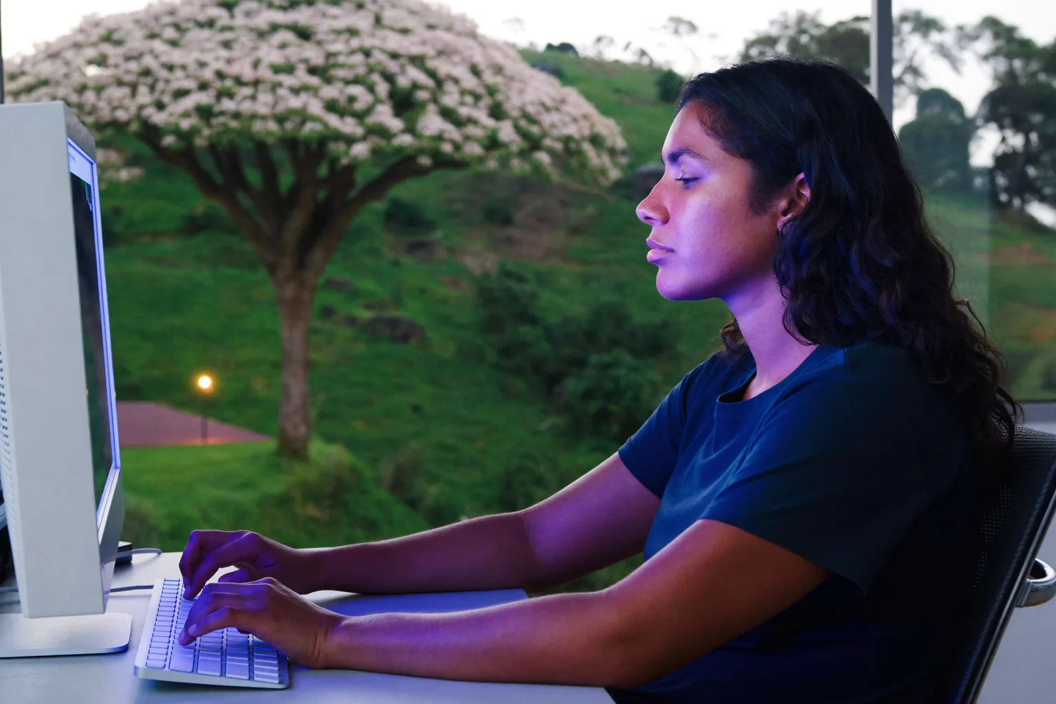 Uma pessoa vista de lado sentada à mesa, com os pés apoiados no chão, as costas contra o apoio da cadeira, os antebraços na altura da mesa e o monitor na altura dos olhos, a postura de referência na mesa