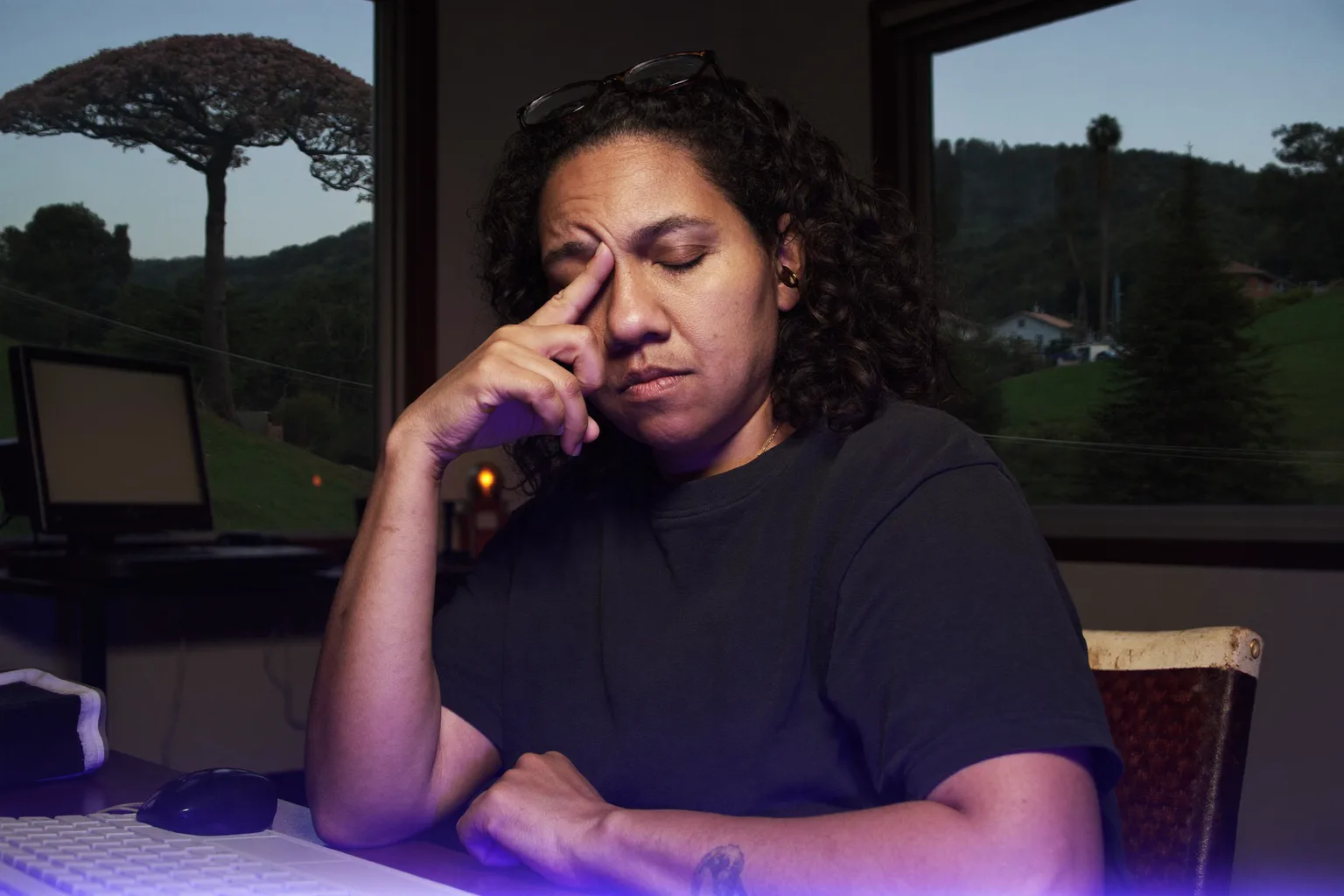 Uma mulher sentada à mesa, em um quarto escuro, de olhos fechados e os óculos empurrados para a testa, apertando a ponte do nariz atrás do monitor, o rosto iluminado por uma luz fria e azulada vinda da tela