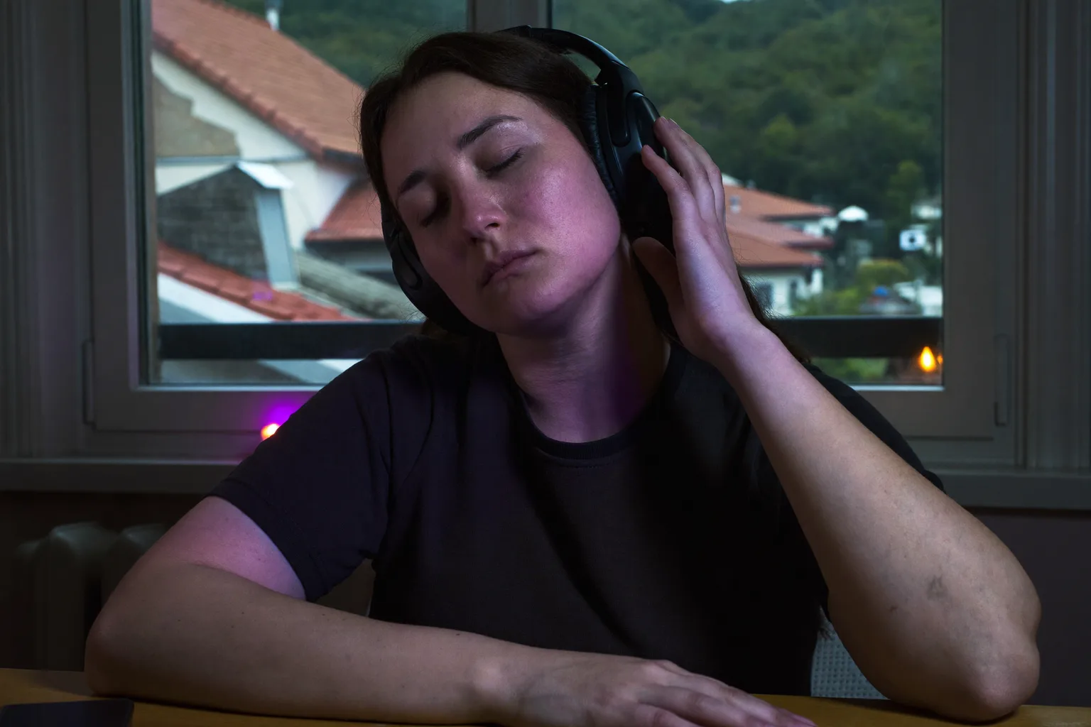 O persoană la birou, purtând căști over-ear, cu ochii închiși și o mână sprijinită pe una dintre căști, cufundată într-o sesiune de lucru cu muzică
