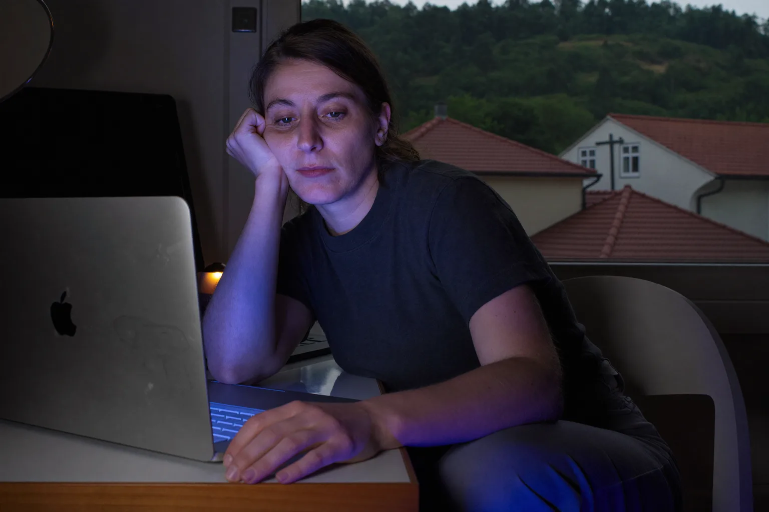O femeie obosită în fața laptopului, târziu în timpul zilei, care se sprijină cu capul în mână, cu ochii grei, uscați și cearcăne, într-o cameră slab luminată