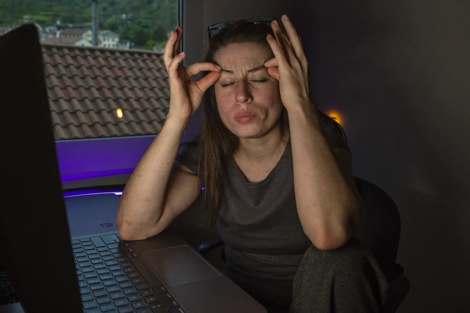 O femeie la birou într-o cameră întunecată, cu ochii închiși și ochelarii ridicați pe frunte, ținându-se de rădăcina nasului deasupra spatelui monitorului, cu fața luminată de albastrul rece al ecranului
