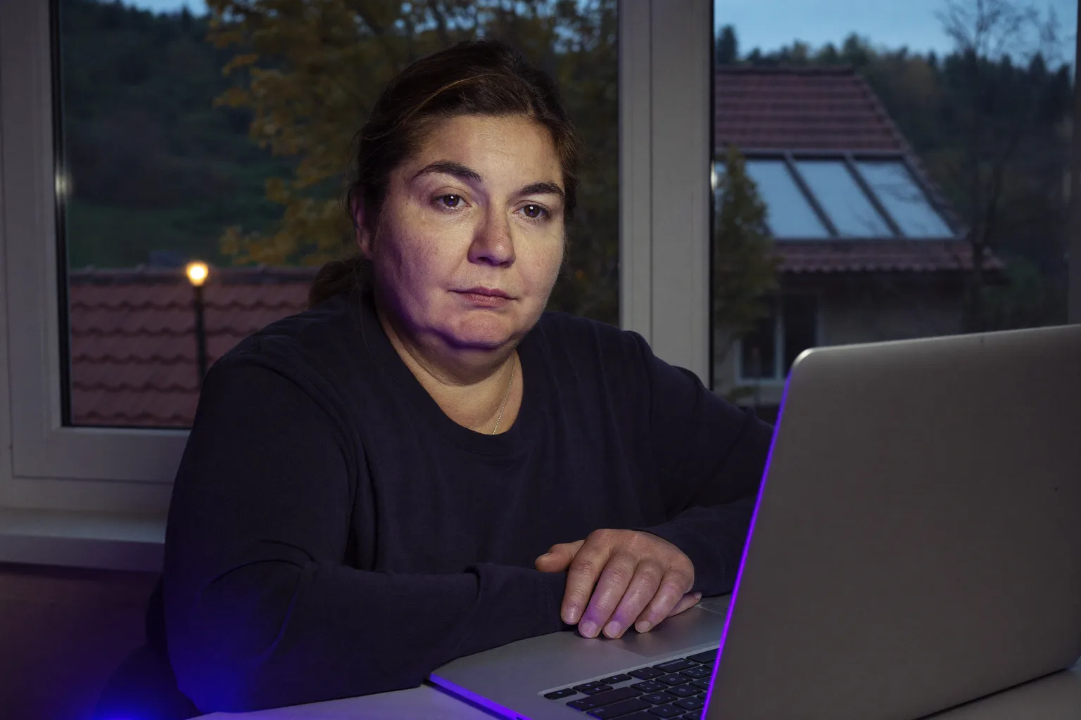 O persoană așezată la un birou de acasă, seara târziu, privind fix, dincolo de ecranul laptopului, cu mâinile încă pe tastatură, arătând epuizarea aplatizată a burnout-ului la locul de muncă