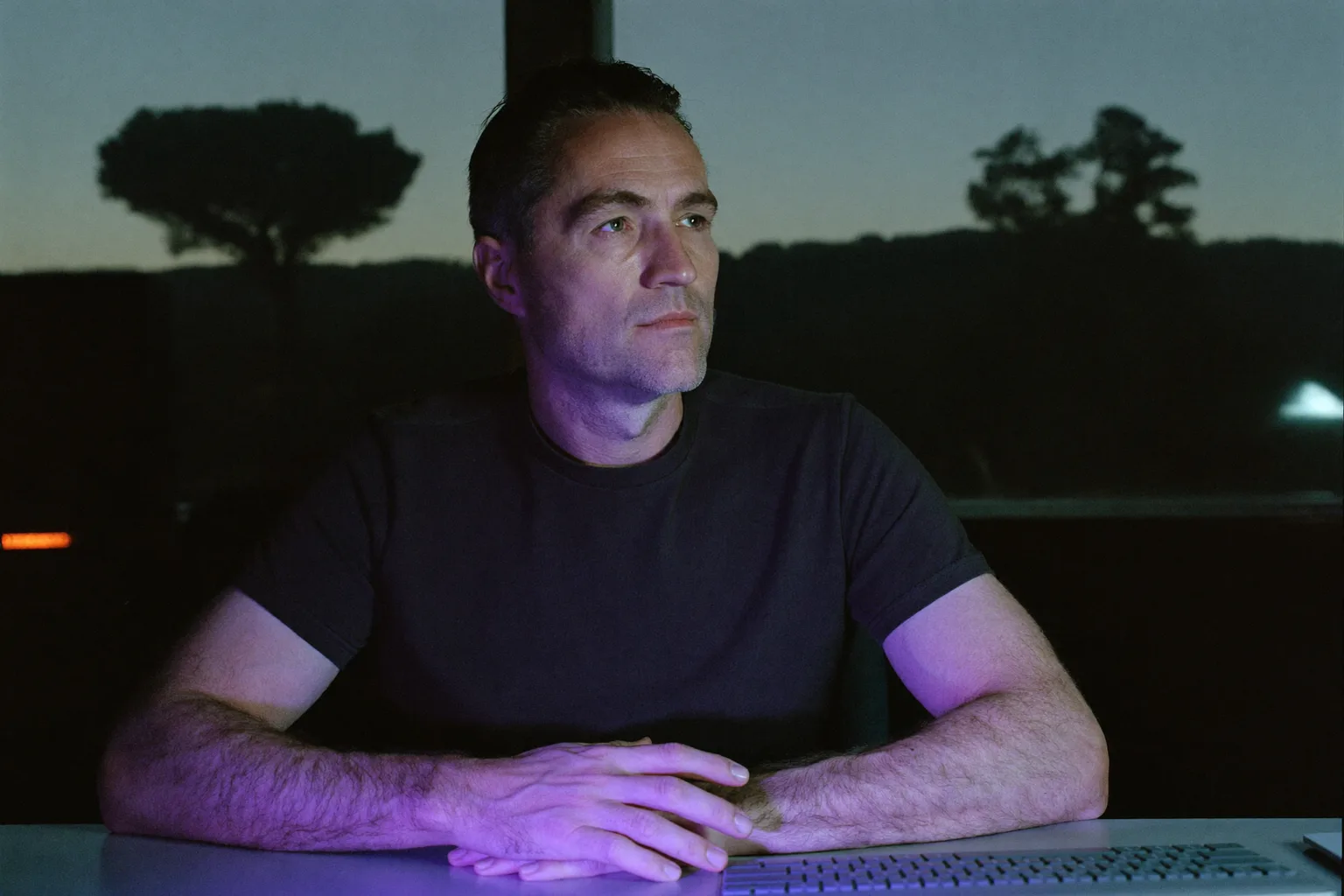 A person sitting at a home desk late in the evening, staring past the laptop screen with their hands still on the keyboard, showing the flat exhaustion of burnout at work