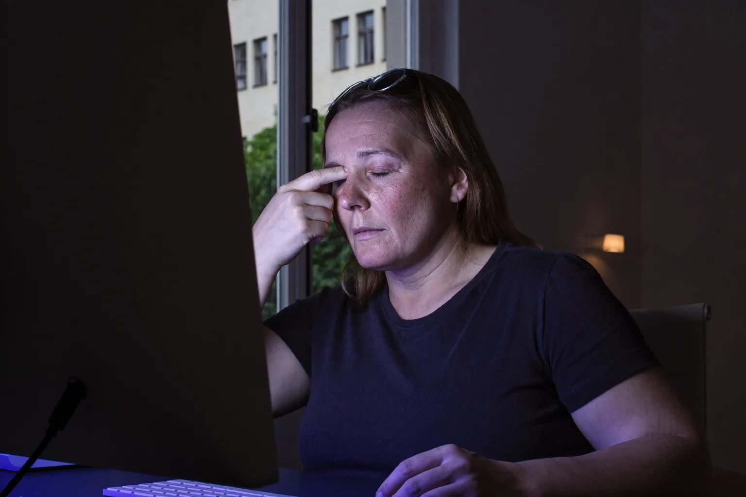 Žena za stolom v tmavej miestnosti, so zavretými očami a okuliarmi vyloženými na čele, si tlačí prsty na koreň nosa nad chrbtom monitora, jej tvár osvetľuje studené modré svetlo obrazovky