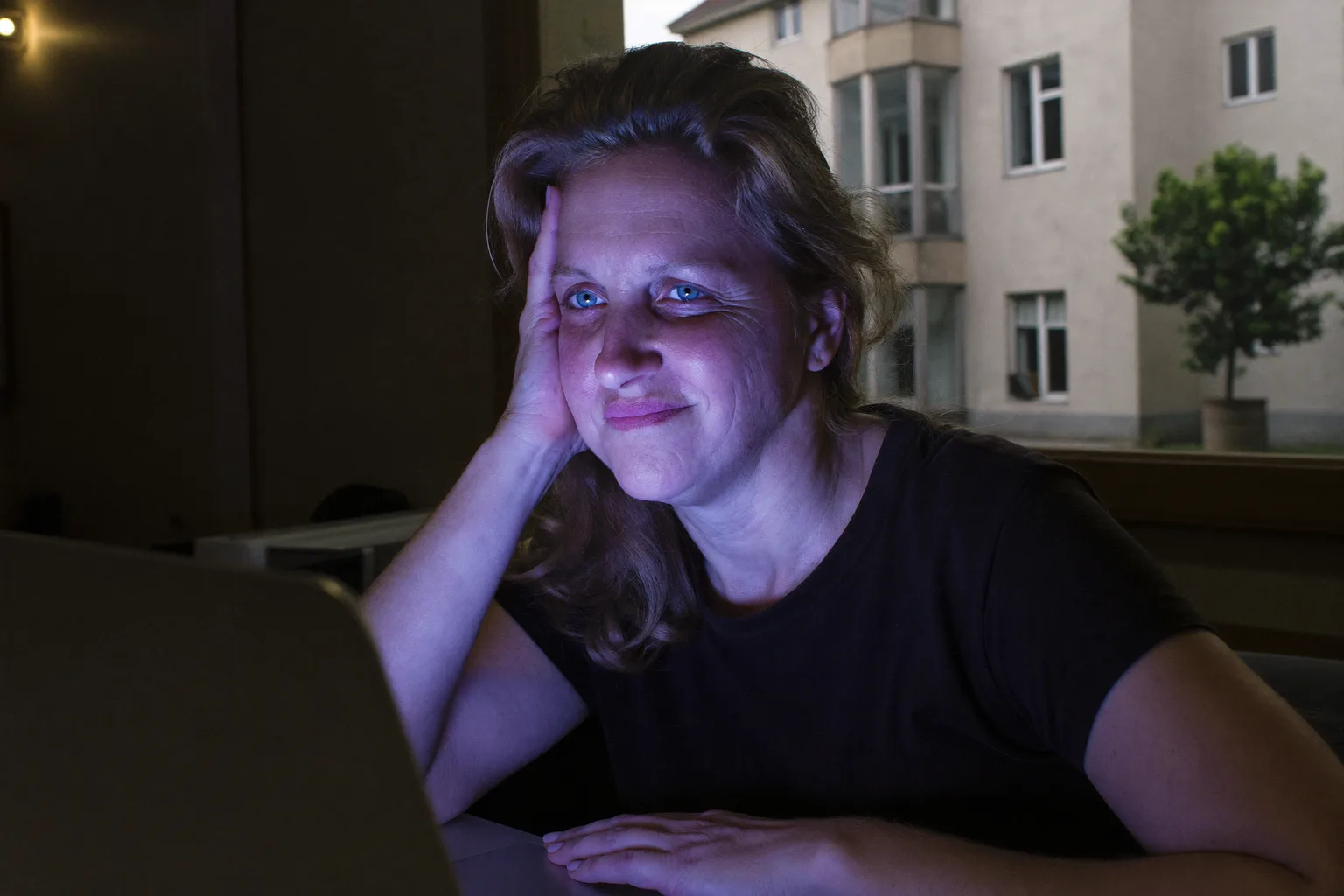 Unavená žena za notebookom neskoro večer, hlavu si opiera o ruku, s ťažkými, suchými očami a tmavými kruhmi v slabo osvetlenej miestnosti
