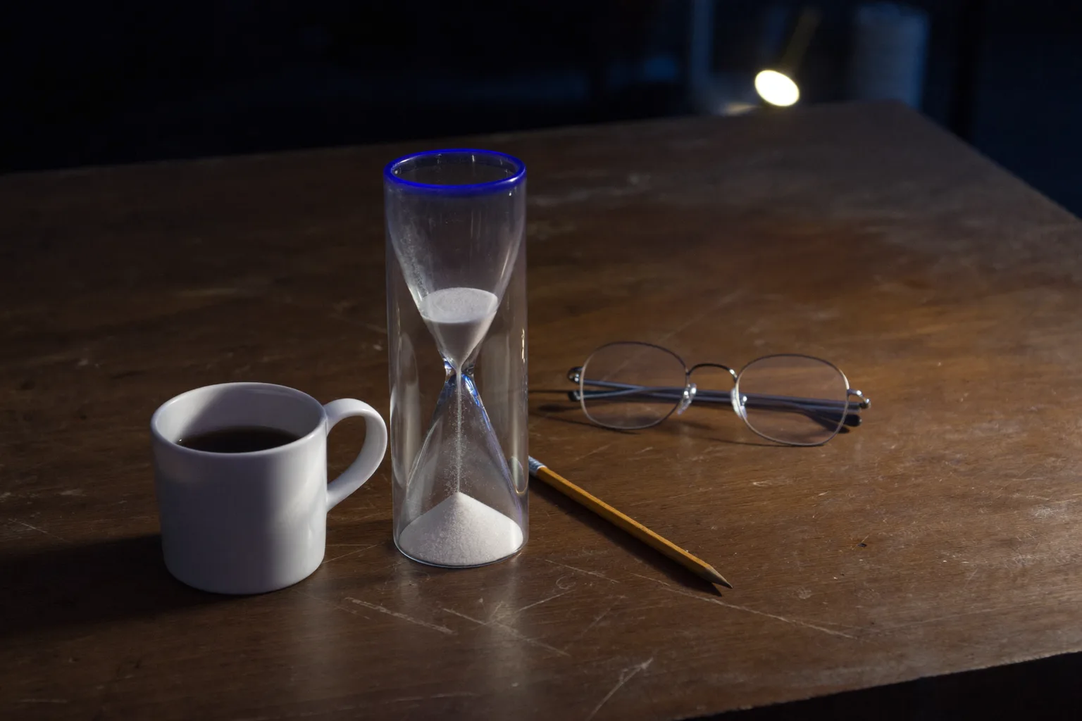An hourglass with the sand still falling, standing on a bare wooden desk at night beside a mug of black coffee, a pencil and a folded pair of reading glasses