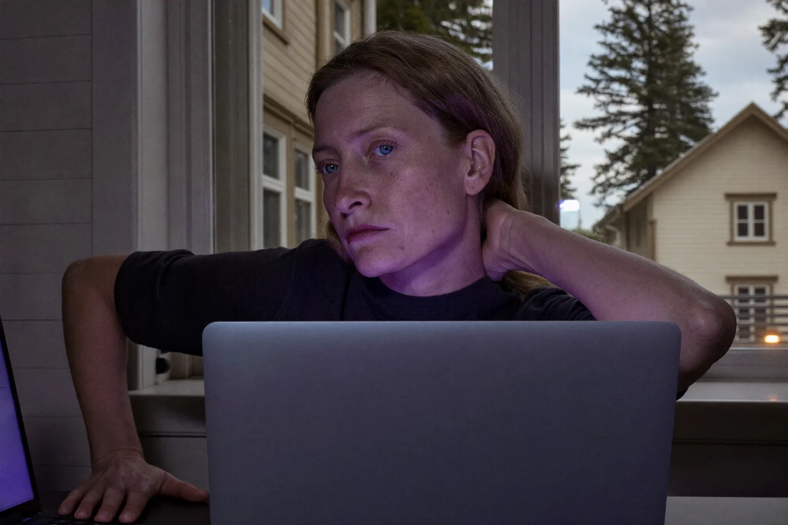 En person vid ett skrivbord som sträcker sig bakåt för att gnugga nedre delen av nacken framför en laptop, vilket visar den stela, värkande nacken efter en lång dag vid datorn