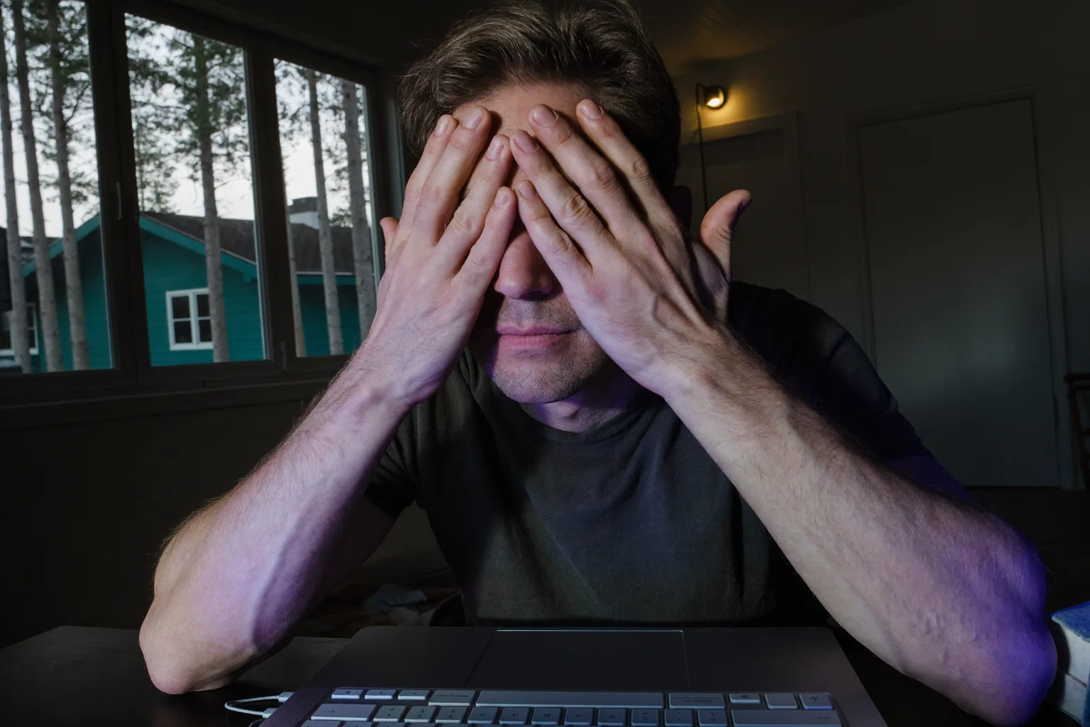 En man vid ett skrivbord i ett svagt upplyst rum trycker handflatorna mot sina slutna ögon för att lindra trötta ögon, ansiktet upplyst av datorskärmen