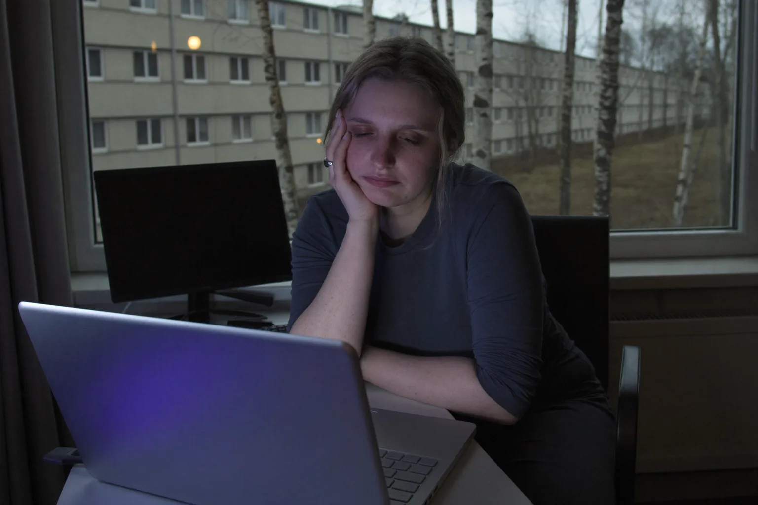 Втомлена жінка за ноутбуком наприкінці дня, підпирає голову рукою, з важкими сухими очима і темними колами в напівтемній кімнаті