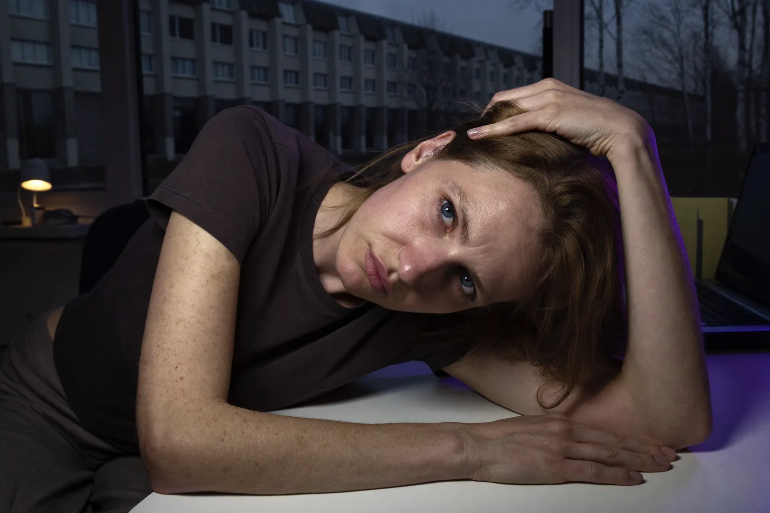 Людина за домашнім столом пізно ввечері, голова схилена на одну руку, пальці в волоссі, погляд опущений і розфокусований, друга рука нерухомо лежить на порожньому столі