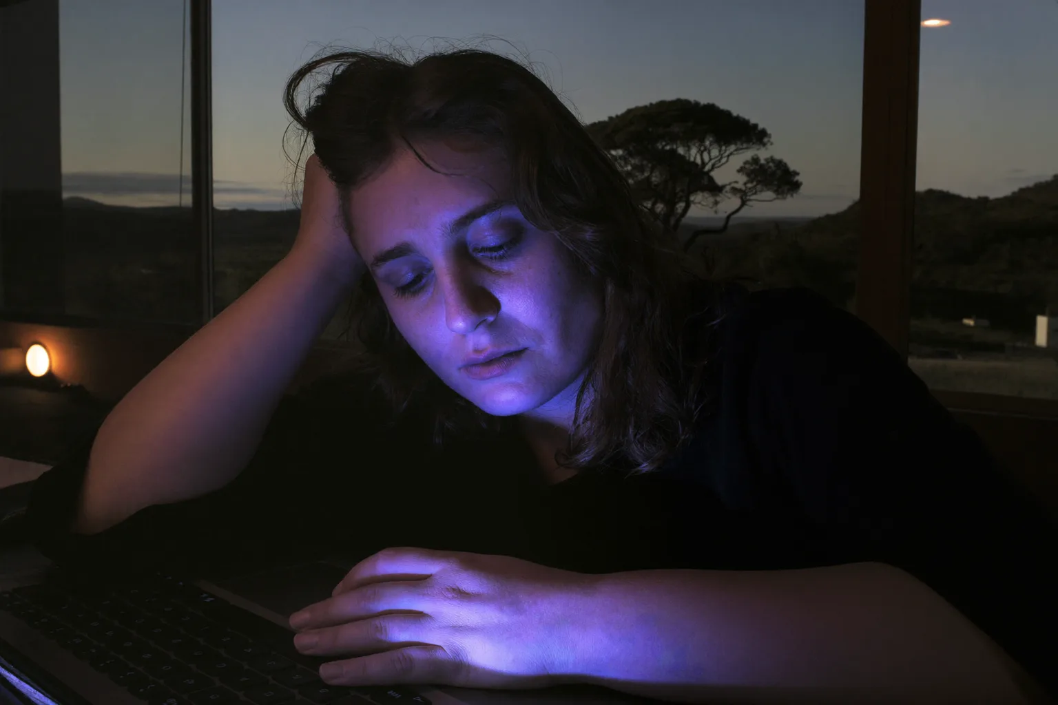 A tired woman at a laptop late in the day, resting her head on her hand, with heavy, dry eyes and dark circles in a dimly lit room