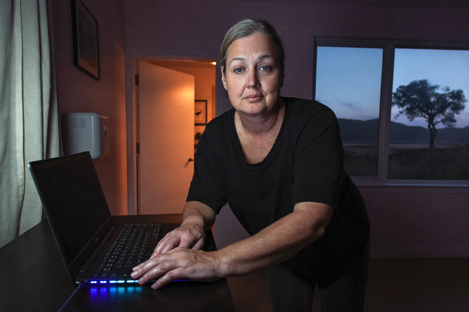 A laptop closed on a home desk in the evening with the room light on and the person walking away, showing the end of a remote working day