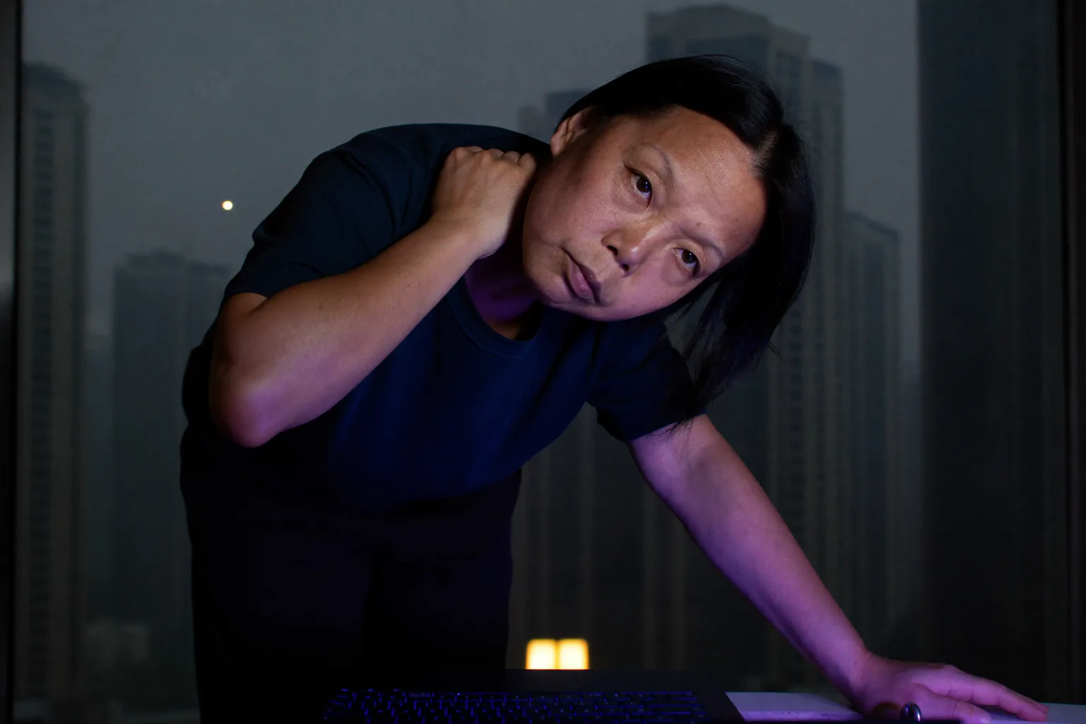 一个人坐在书桌前，在笔记本电脑旁边伸手揉着脖子根部，展现出长时间用电脑后又僵又酸的脖子