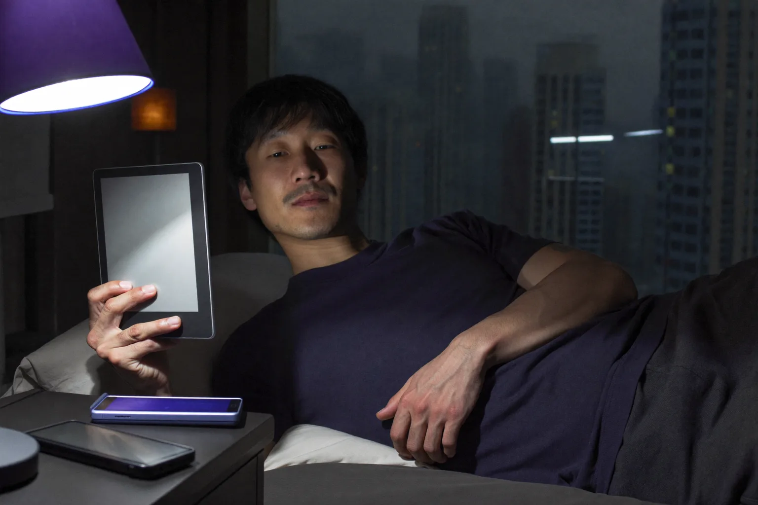 深夜，一名男子躺在床上用电子书阅读器看书，页面透出的微光几乎照不到脸上，而床头柜上一部屏幕朝上的手机，把旁边那盏灯罩染成一片生硬冰冷的蓝光