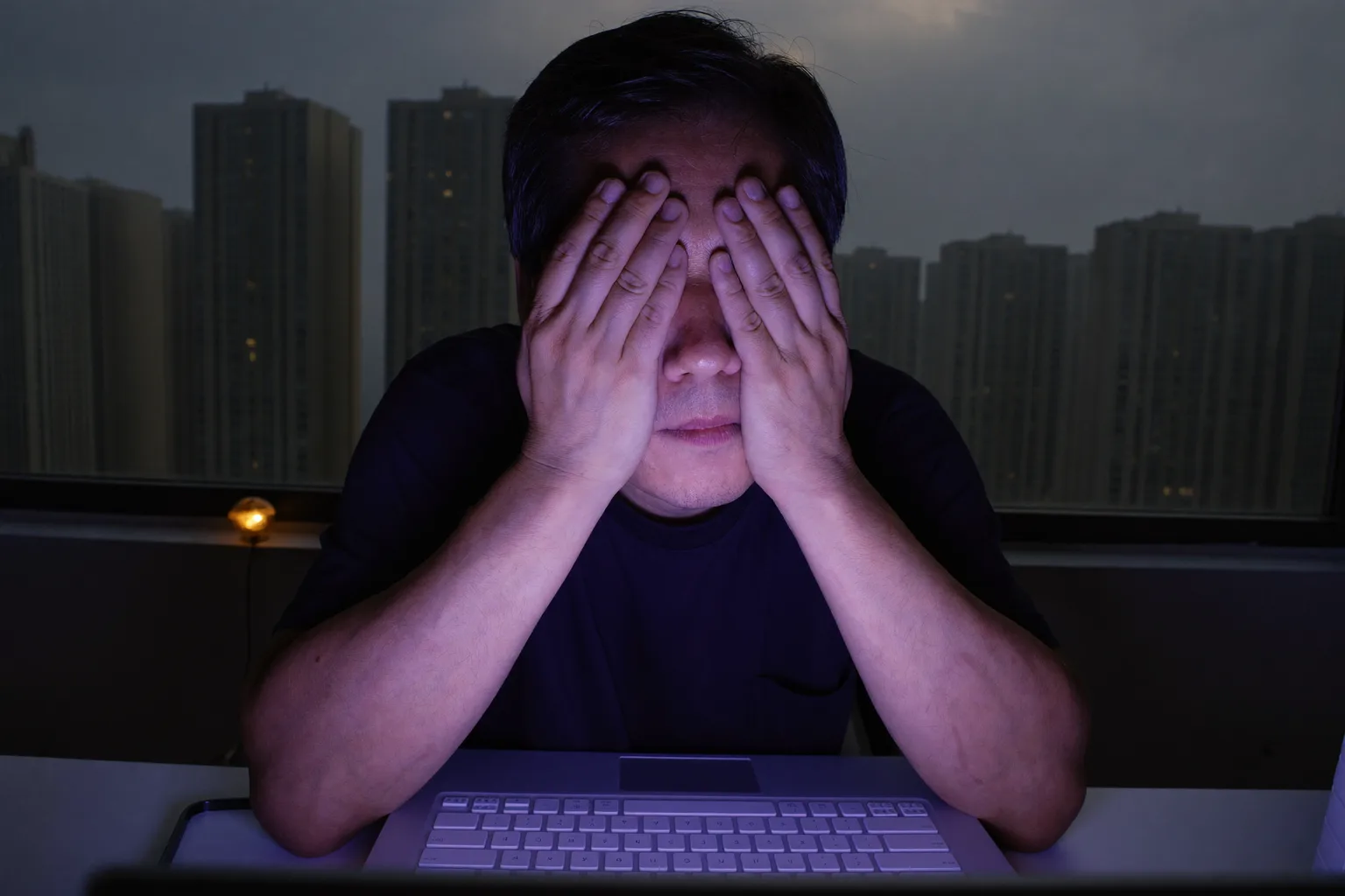 一名男子坐在昏暗房間的桌前，用掌心捂住閉著的雙眼來舒緩眼睛疲勞，臉龐被筆電螢幕的光線照亮
