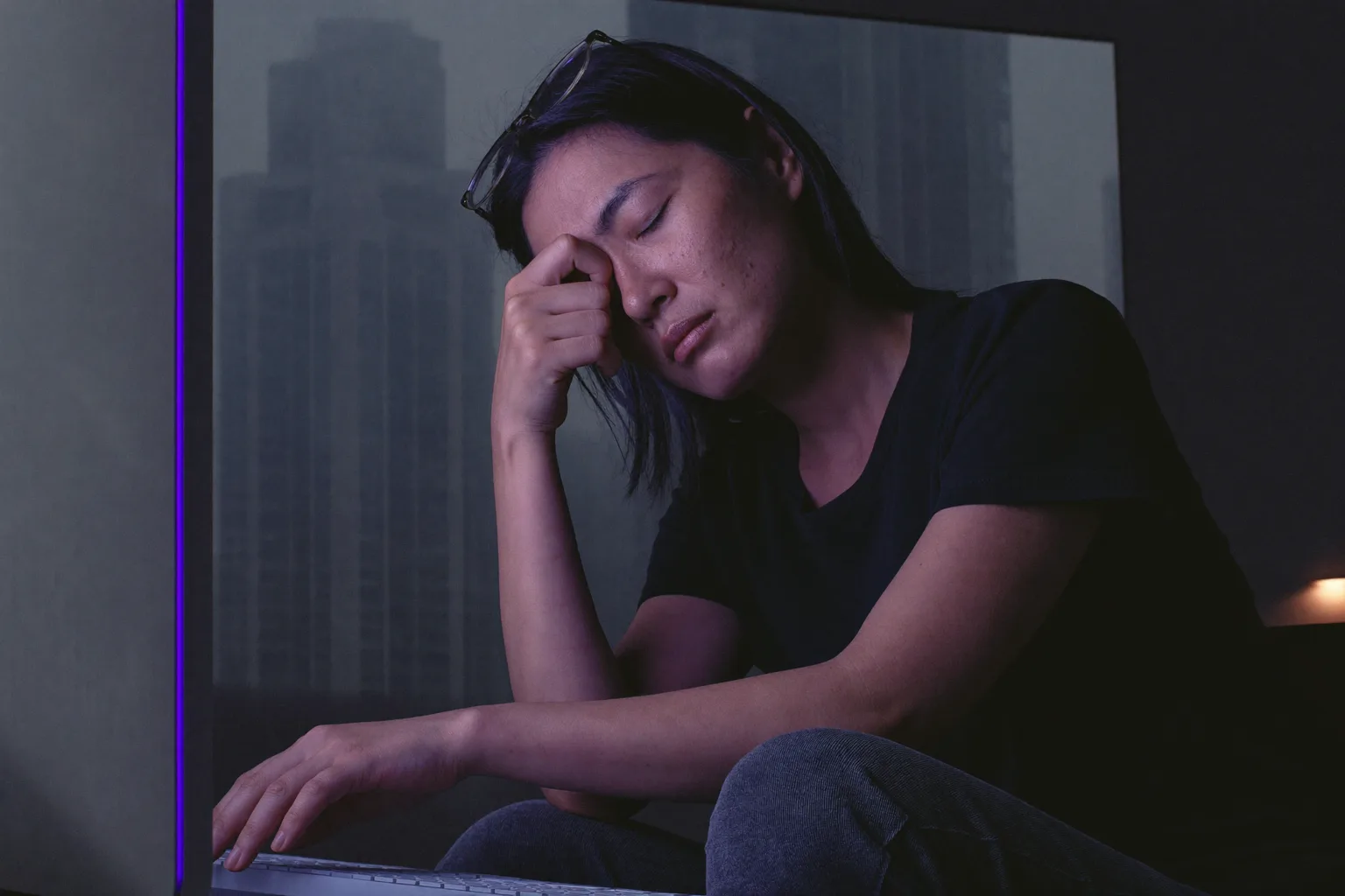 一位女性坐在昏暗房間的辦公桌前，閉著眼睛，眼鏡推到額頭上，手指捏著鼻樑，臉被螢幕的冷藍光照亮，螢幕背面就在她面前